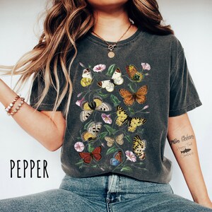 May include: A dark grey t-shirt with a vintage-style print of colourful butterflies and flowers. The shirt has a crew neck and short sleeves.