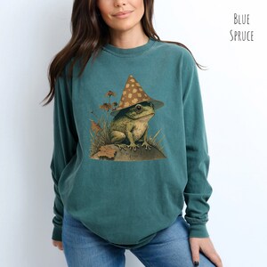 Pode incluir: Uma camiseta de manga comprida azul-esverdeada com um gr&aacute;fico de um sapo usando um chap&eacute;u marrom e branco com manchas. O sapo est&aacute; sentado em uma rocha com grama verde e folhas ao redor.