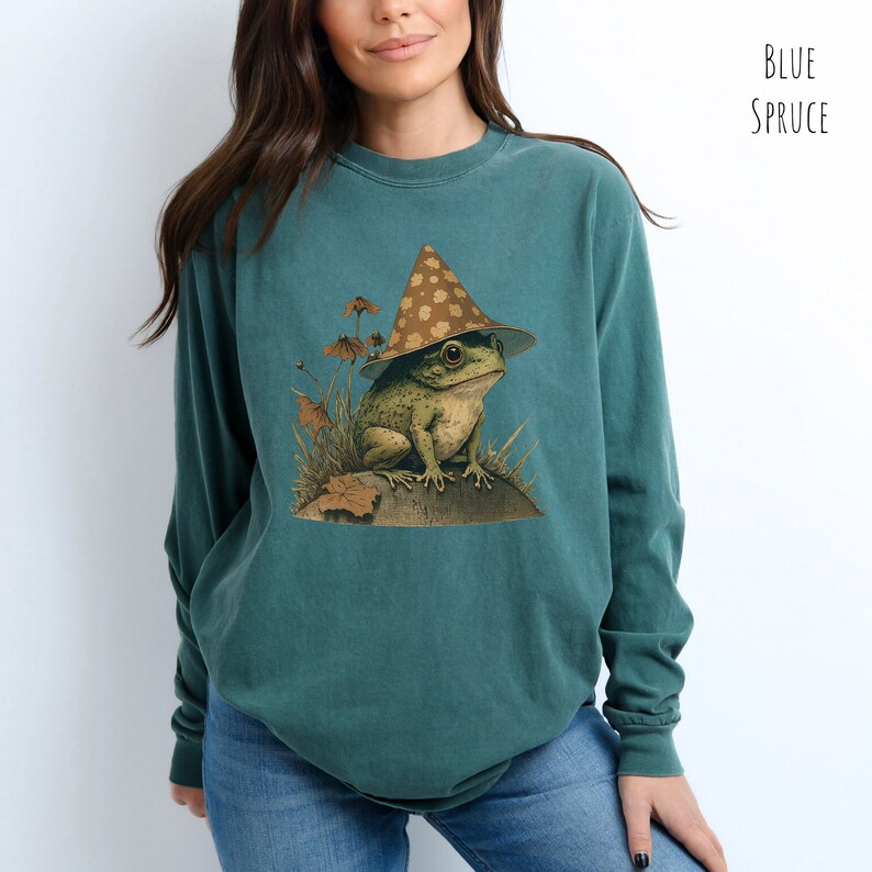 Pode incluir: Uma camiseta de manga comprida azul-esverdeada com um gr&aacute;fico de um sapo usando um chap&eacute;u marrom e branco com manchas. O sapo est&aacute; sentado em uma rocha com grama verde e folhas ao redor.