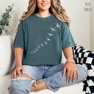 May include: A teal blue t-shirt with a white dragonfly design. The dragonflies are arranged in a line, flying upwards. The shirt is made of a soft, comfortable material and has a relaxed fit.