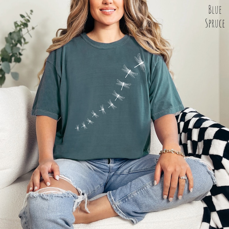 May include: A teal blue t-shirt with a white dragonfly design. The dragonflies are arranged in a line, flying upwards. The shirt is made of a soft, comfortable material and has a relaxed fit.