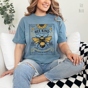 K&ouml;nnte beinhalten: Ein hellblaues T-Shirt mit einer Grafik im Vintage-Stil, die eine Biene und eine Sonnenblume zeigt. Die Grafik lautet "Bee Kind, Bee Positive, Bee Humble, Bee Honest, Bee Grateful, Believe in Yourself".
