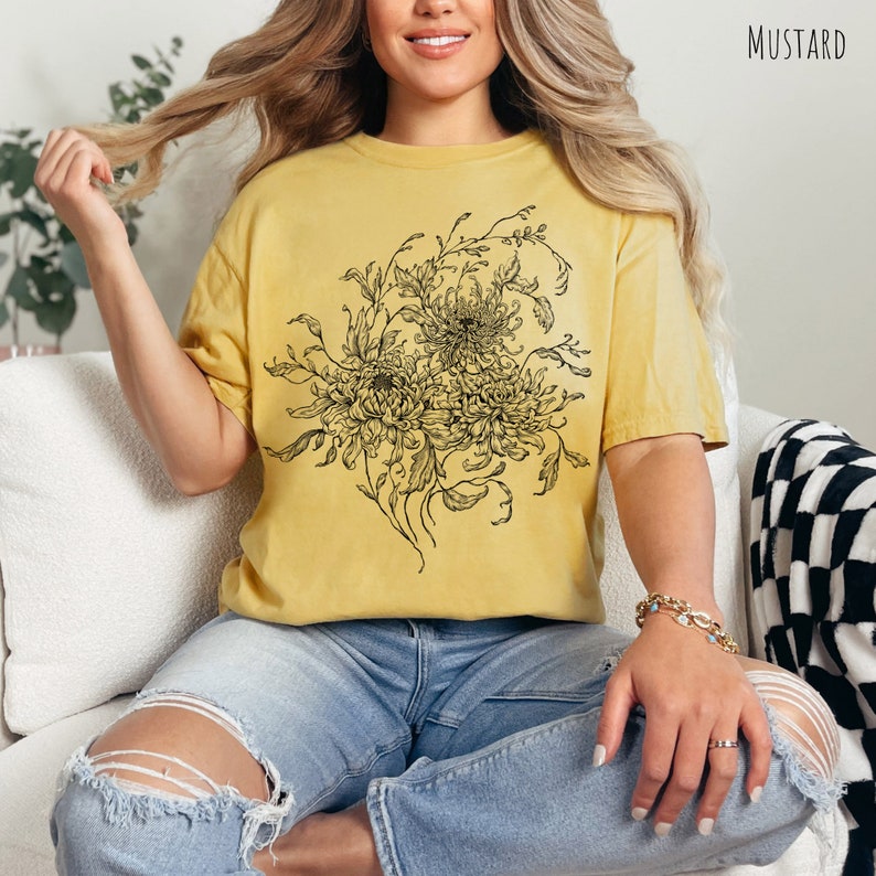 May include: A mustard yellow t-shirt featuring a black and white floral design. The design depicts a bouquet of chrysanthemums.