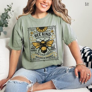 K&ouml;nnte beinhalten: Ein gr&uuml;nes T-Shirt mit einer Grafik im Vintage-Stil, die eine Biene und eine Sonnenblume zeigt. Die Grafik lautet "Bee Kind", "Bee Positive", "Bee Humble", "Bee Honest", "Bee Grateful" und "Believe in Yourself".