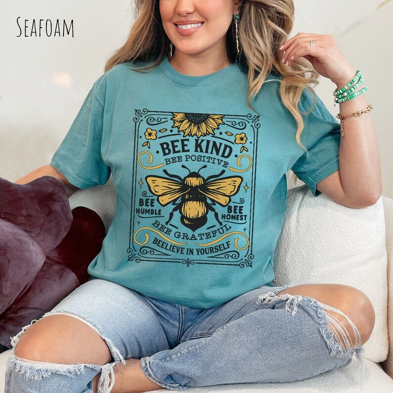 K&ouml;nnte beinhalten: Ein t&uuml;rkisfarbenes T-Shirt mit einer Vintage-Grafik einer Biene und einer Sonnenblume. Die Grafik lautet "Bee Kind, Bee Positive, Bee Humble, Bee Honest, Bee Grateful, Believe in Yourself".