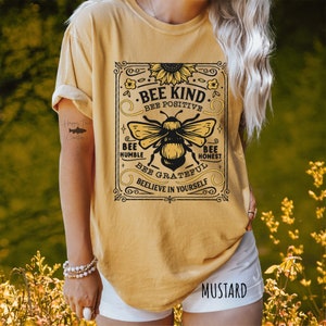 K&ouml;nnte beinhalten: Ein senfgelbes T-Shirt mit einer Grafik im Vintage-Stil, die eine Biene und den Text "Bee Kind. Bee Positive. Bee Humble. Bee Honest. Bee Grateful. Believe in Yourself." zeigt.