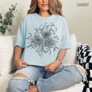May include: A light blue t-shirt featuring a black and white floral design. The design depicts a bouquet of flowers with intricate details. The shirt is made from a soft, comfortable fabric and has a relaxed fit.