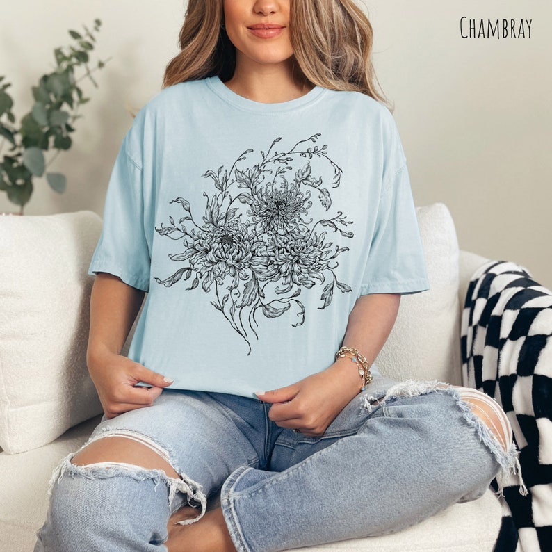 May include: A light blue t-shirt featuring a black and white floral design. The design depicts a bouquet of flowers with intricate details. The shirt is made from a soft, comfortable fabric and has a relaxed fit.