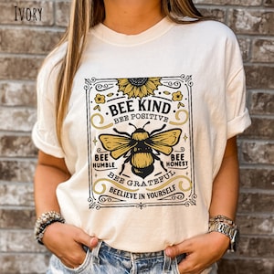 K&ouml;nnte beinhalten: Ein wei&szlig;es T-Shirt mit einer Grafik im Vintage-Stil, die eine Biene und eine Sonnenblume zeigt. Die Grafik lautet "Bee Kind", "Bee Positive", "Bee Humble", "Bee Honest", "Bee Grateful" und "Believe in Yourself".