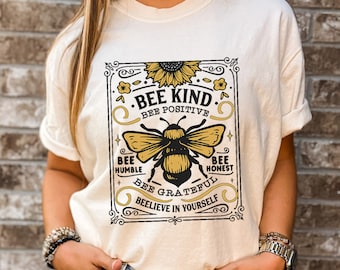 Peut inclure: Un t-shirt blanc avec un graphique de style vintage représentant une abeille et un tournesol. Le graphique dit "Bee Kind", "Bee Positive", "Bee Humble", "Bee Honest", "Bee Grateful", et "Believe in Yourself".
