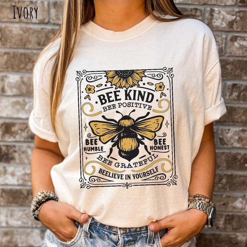 K&ouml;nnte beinhalten: Ein wei&szlig;es T-Shirt mit einer Grafik im Vintage-Stil, die eine Biene und eine Sonnenblume zeigt. Die Grafik lautet "Bee Kind", "Bee Positive", "Bee Humble", "Bee Honest", "Bee Grateful" und "Believe in Yourself".