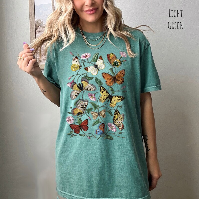 May include: A light green t-shirt featuring a colourful butterfly print. The butterflies are arranged in a cluster on the front of the shirt.
