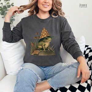Pode incluir: Uma camisa de moletom cinza escura com um gr&aacute;fico de um sapo usando um chap&eacute;u marrom e amarelo com manchas. O sapo est&aacute; sentado em uma pedra com grama verde e folhas ao redor.