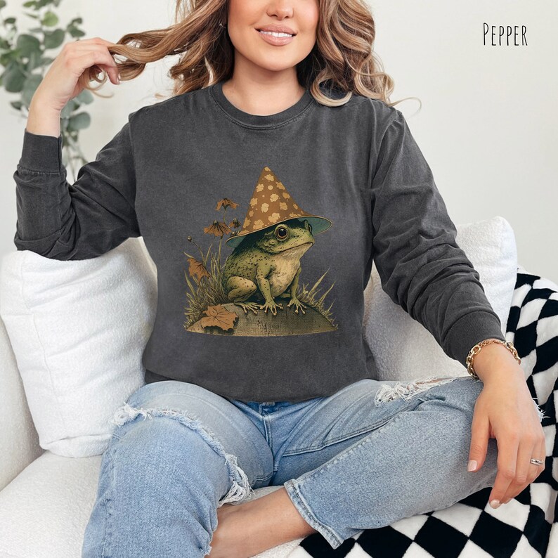 Pode incluir: Uma camisa de moletom cinza escura com um gr&aacute;fico de um sapo usando um chap&eacute;u marrom e amarelo com manchas. O sapo est&aacute; sentado em uma pedra com grama verde e folhas ao redor.
