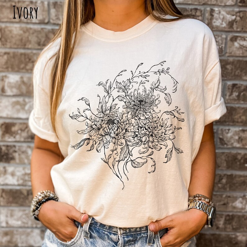 May include: A white t-shirt featuring a black and white floral design. The design depicts a bouquet of chrysanthemums with intricate details.