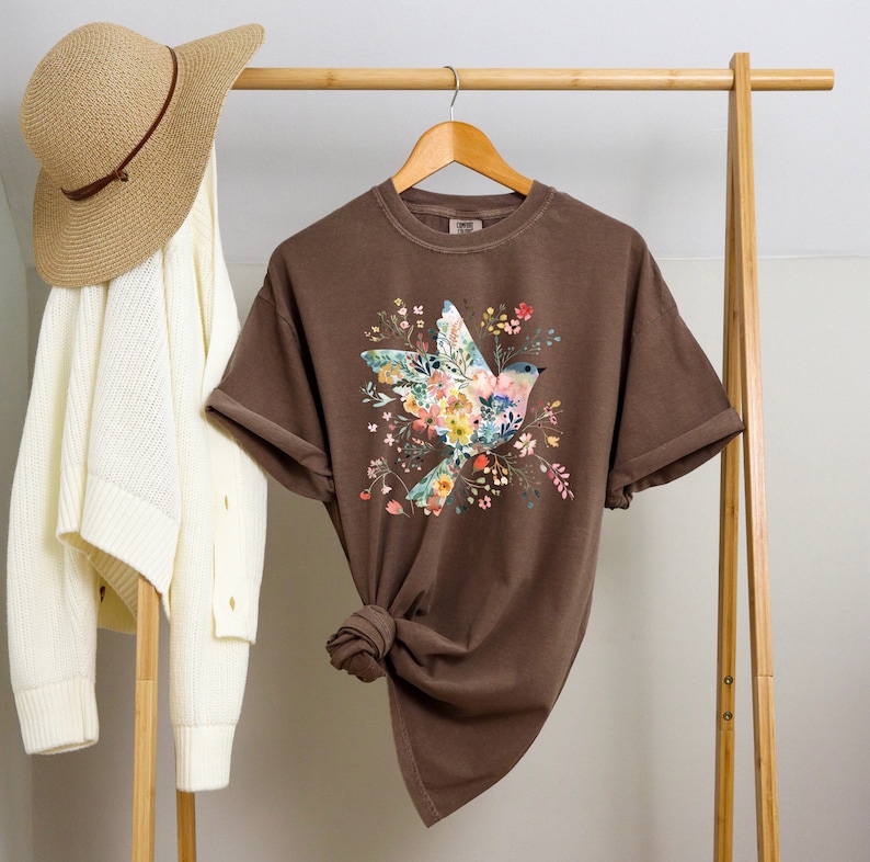 Op de afbeelding: Een bruin T-shirt met een bloemenprint met een vogel. De vogel is gemaakt van bloemen en heeft een blauwe kop en vleugels. Het shirt hangt aan een houten hanger.