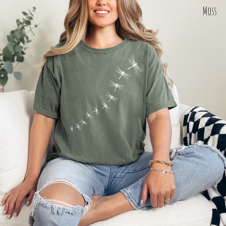 May include: A woman wearing a green t-shirt with a white dragonfly design. The shirt has a relaxed fit and a crew neck. The woman is sitting on a white couch with a black and white checkered throw blanket.