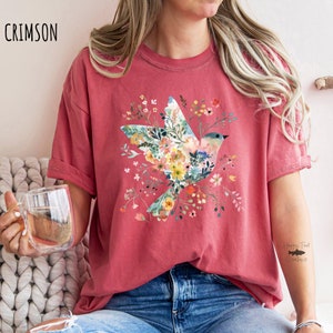 Op de afbeelding: Een crimson t-shirt met een bloemenontwerp van een vogel in vlucht. Het ontwerp bevat een verscheidenheid aan kleuren, waaronder roze, geel, blauw en groen. Het shirt heeft een ontspannen pasvorm en korte mouwen. De tekst "Happy Trout Creations" is zichtbaar op de rechtermouw.