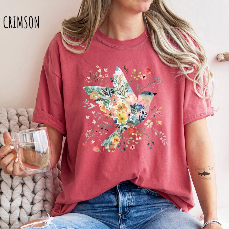 Op de afbeelding: Een crimson t-shirt met een bloemenontwerp van een vogel in vlucht. Het ontwerp bevat een verscheidenheid aan kleuren, waaronder roze, geel, blauw en groen. Het shirt heeft een ontspannen pasvorm en korte mouwen. De tekst "Happy Trout Creations" is zichtbaar op de rechtermouw.