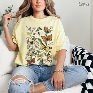 May include: A yellow t-shirt featuring a vintage-style print of butterflies and flowers. The butterflies are in various colours, including orange, brown, yellow, and blue.