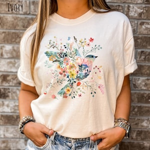 Op de afbeelding: Een wit T-shirt met een bloemenprint met een blauwe vogel met roze en gele accenten. Het ontwerp is gecentreerd op het shirt en is omringd door verschillende bloemen in tinten roze, geel en blauw.