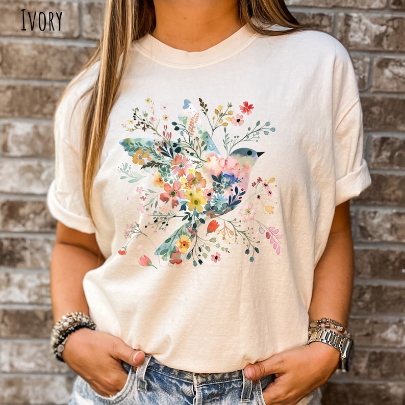 Op de afbeelding: Een wit T-shirt met een bloemenprint met een blauwe vogel met roze en gele accenten. Het ontwerp is gecentreerd op het shirt en is omringd door verschillende bloemen in tinten roze, geel en blauw.
