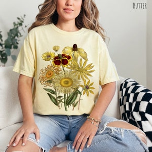 Op de afbeelding: Een geel T-shirt met een vintage bloemenprint met gele, rode en bruine bloemen. De bloemen zijn in een boeket gerangschikt en omgeven door groene bladeren.
