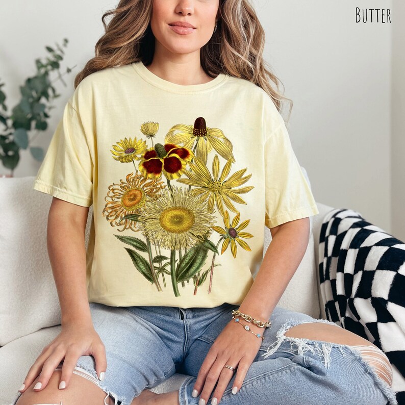 Op de afbeelding: Een geel T-shirt met een vintage bloemenprint met gele, rode en bruine bloemen. De bloemen zijn in een boeket gerangschikt en omgeven door groene bladeren.