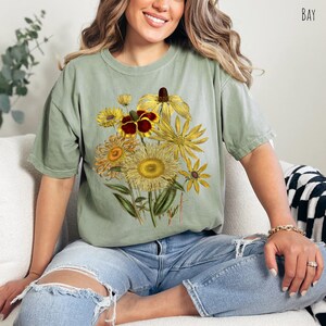 Op de afbeelding: Een saliegroen T-shirt met een vintage bloemenprint met gele, oranje en bruine bloemen. De bloemen zijn in een boeket gerangschikt en hebben een botanische illustratiestijl.