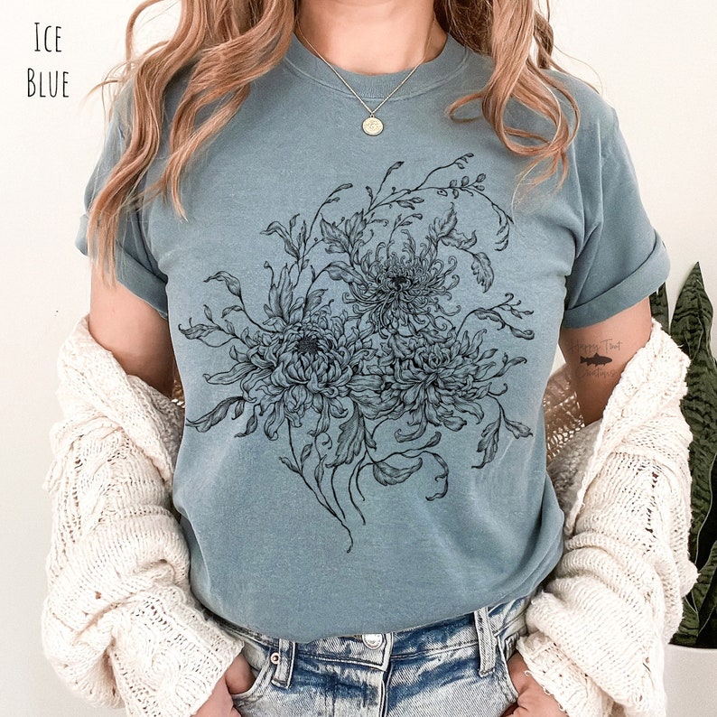 May include: A light blue t-shirt with a black and white floral design featuring chrysanthemums. The design is centred on the shirt and covers most of the front.