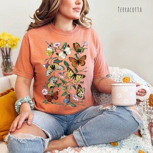 May include: A terracotta coloured t-shirt featuring a graphic of colourful butterflies and flowers. The word "TERRACOTTA" is printed in white in the top right corner of the image.