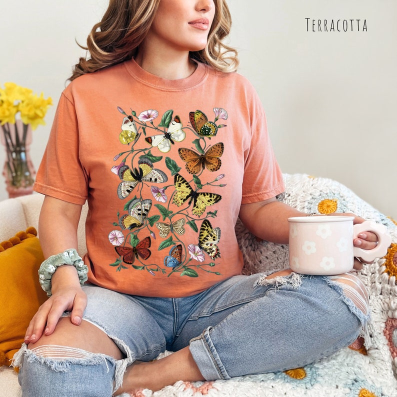 May include: A terracotta coloured t-shirt featuring a graphic of colourful butterflies and flowers. The word "TERRACOTTA" is printed in white in the top right corner of the image.