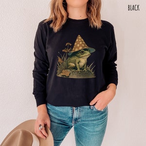 Pode incluir: Uma camisa preta de manga comprida com um gr&aacute;fico de um sapo usando um chap&eacute;u em forma de cogumelo. O sapo est&aacute; sentado em uma rocha com folhagem verde ao redor.