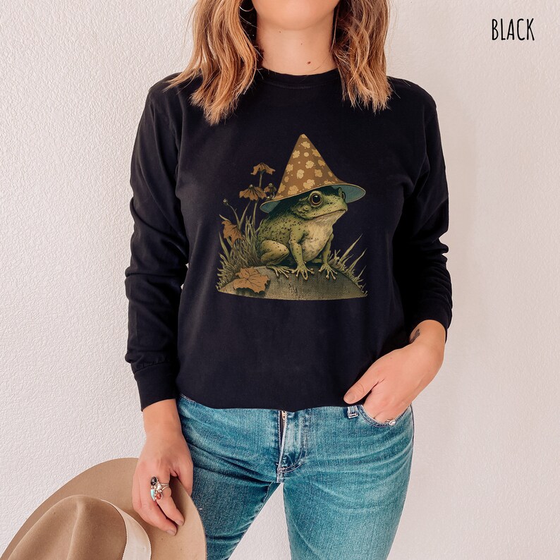Pode incluir: Uma camisa preta de manga comprida com um gr&aacute;fico de um sapo usando um chap&eacute;u em forma de cogumelo. O sapo est&aacute; sentado em uma rocha com folhagem verde ao redor.