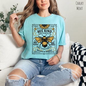 K&ouml;nnte beinhalten: Ein hellblaues T-Shirt mit einer Grafik im Vintage-Stil, die eine Biene und den Text "Bee Kind. Bee Positive. Bee Humble. Bee Honest. Bee Grateful. Believe in Yourself." zeigt.