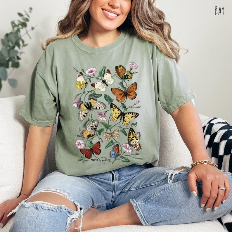 May include: A green t-shirt featuring a vintage-style print of butterflies and flowers. The butterflies are in various colours, including orange, yellow, blue, and red.