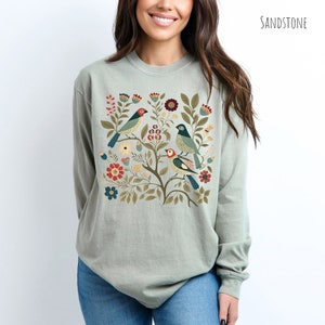 May include: A light green long-sleeved t-shirt with a colorful floral design featuring birds. The design includes red, blue, green, and yellow flowers and birds.