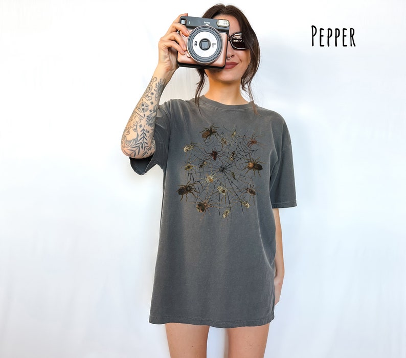 May include: An oversized t-shirt in a dark grey colour with a graphic print of a spider web with spiders in the centre. The text "PEPPER" is in the top right corner of the image.