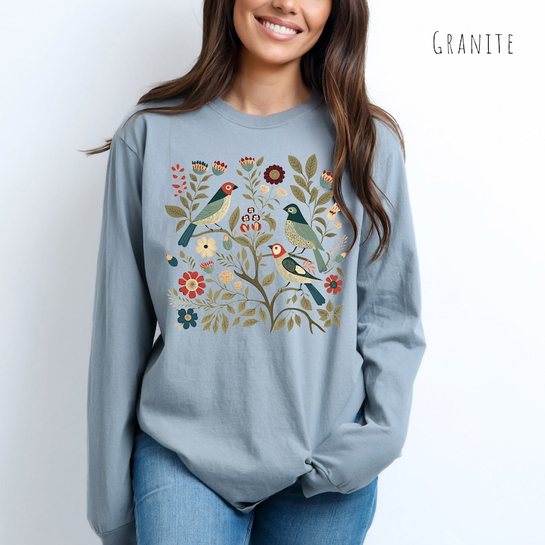 May include: A light blue long-sleeved t-shirt with a floral design featuring three birds. The birds are green, blue, and red with yellow beaks. The flowers are red, yellow, and green.