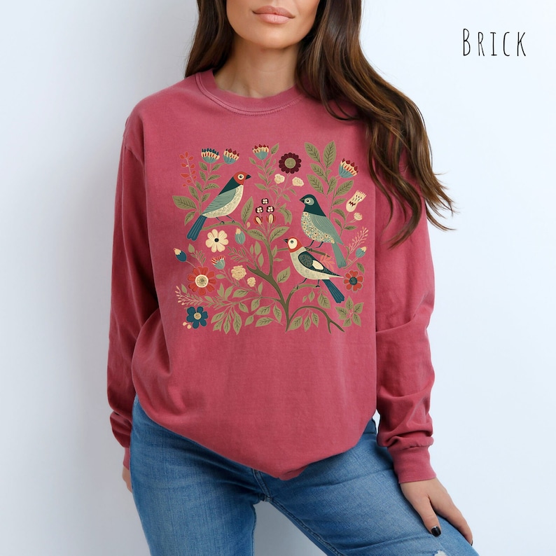 May include: A red long-sleeved sweatshirt with a floral and bird design. The design features three birds with blue, green, and yellow accents, surrounded by flowers and leaves.