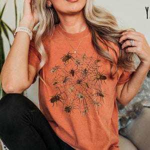 May include: A woman wearing a burnt orange t-shirt with a graphic of a spider web and spiders. The text "YAM" is in the top right corner of the image.