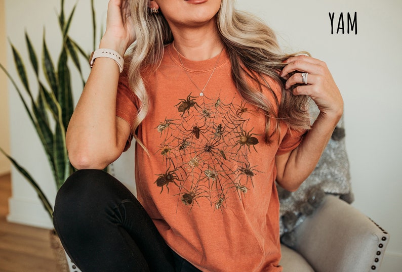 May include: A woman wearing a burnt orange t-shirt with a graphic of a spider web and spiders. The text "YAM" is in the top right corner of the image.