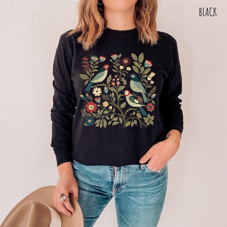 May include: Black long-sleeved shirt with a colorful floral and bird design. The design features a tree with flowers, leaves, and birds in shades of blue, green, red, and yellow.