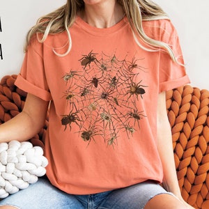 May include: A terracotta orange t-shirt featuring a graphic print of a spider web with spiders.