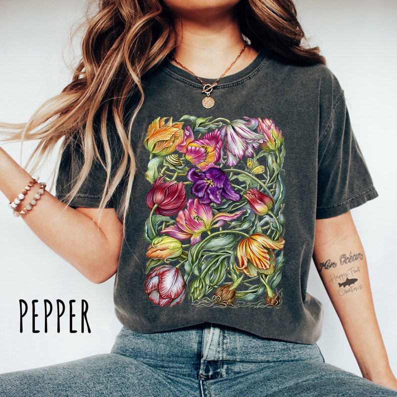 Op de afbeelding: Een donkergrijs T-shirt met een kleurrijke bloemenprint van tulpen. De print bevat een verscheidenheid aan kleuren, waaronder rood, roze, paars, geel en groen. Het shirt heeft een ronde hals en korte mouwen. De tekst "More Dreams Happy That Creations" is gedrukt op de rechtermouw van het shirt.