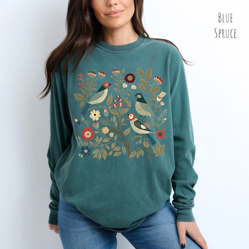 May include: A teal blue long-sleeved t-shirt with a colorful floral and bird design. The design features four birds with red, blue, and yellow accents, surrounded by green leaves and flowers.
