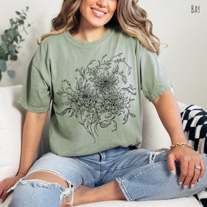 May include: A green t-shirt featuring a black and white floral design. The design depicts a bouquet of flowers with intricate details.