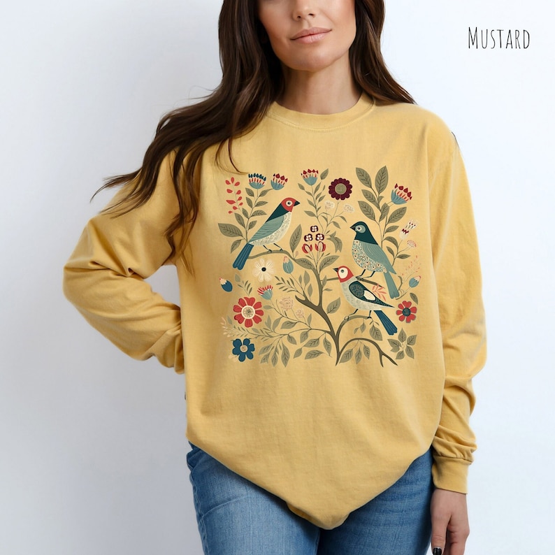 May include: A mustard yellow long-sleeved shirt with a floral and bird design. The design features three birds with blue, green, and red accents, surrounded by green leaves and red, blue, and white flowers.