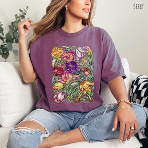 Op de afbeelding: Een paars T-shirt met een kleurrijke bloemenprint van tulpen. De print bevat een verscheidenheid aan kleuren, waaronder rood, geel, oranje, roze en paars. De bloemen zijn in een cluster gerangschikt en worden omringd door groene bladeren.