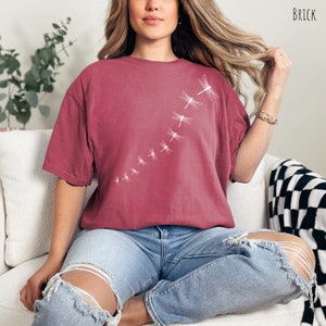 May include: A maroon t-shirt with a white dragonfly design. The dragonflies are arranged in a diagonal line, getting smaller as they go up the shirt.